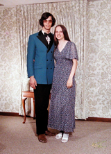 Gary and his girlfriend posing at her prom, he in a blue tuxedo with thick sideburns, 70s style long hair, and pink tinted glasses.